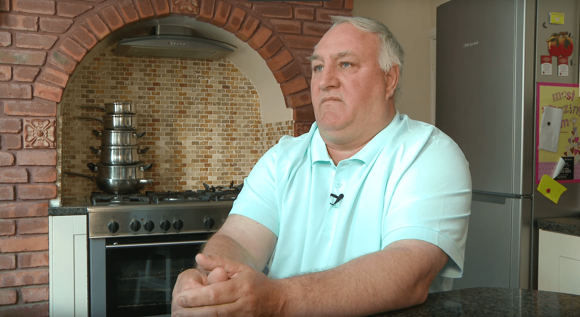 A man sits in his kitchen clasping his hands, his expression is serious