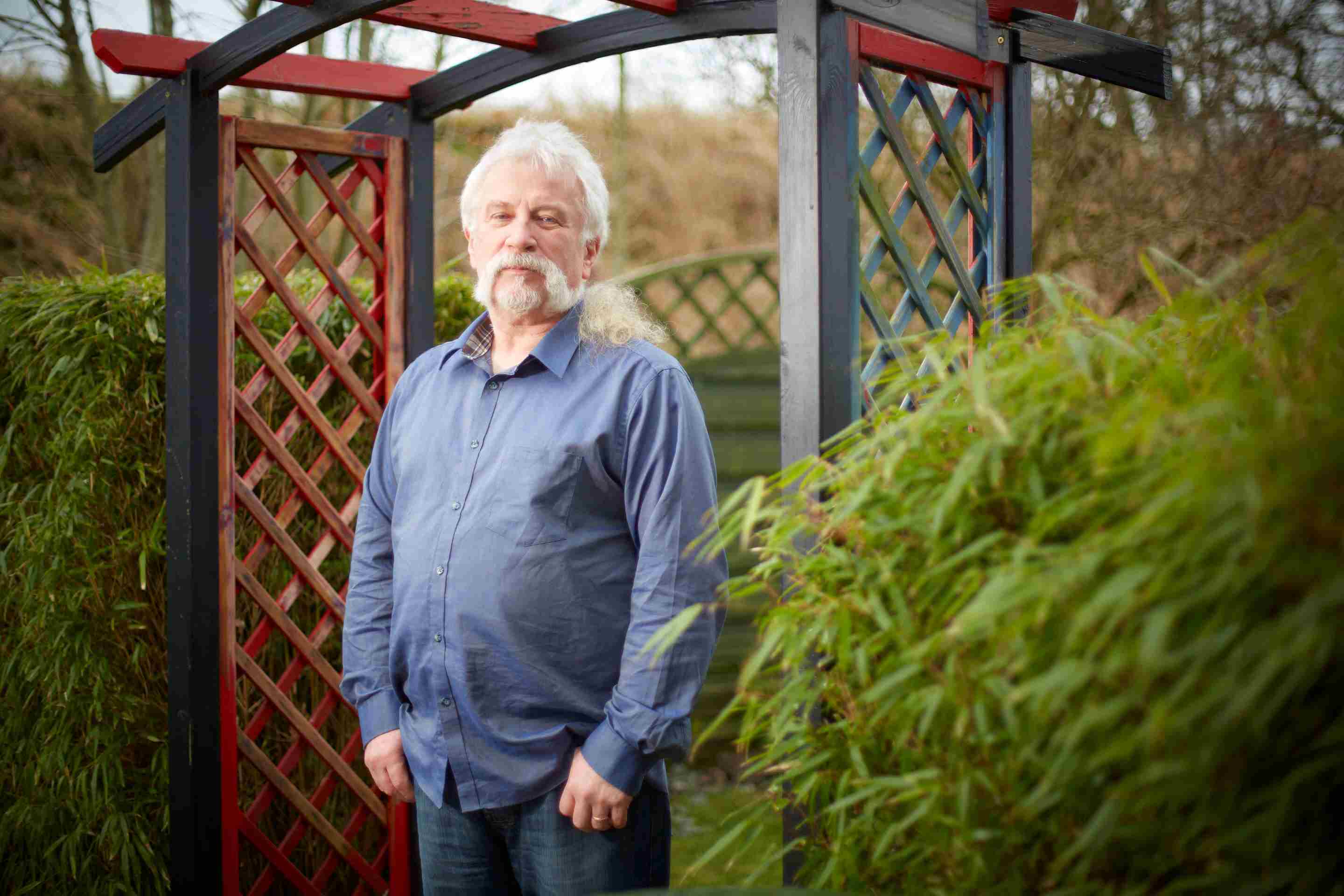 A man in a blue shirt with white hair stands in a garden archway, hands in pockets