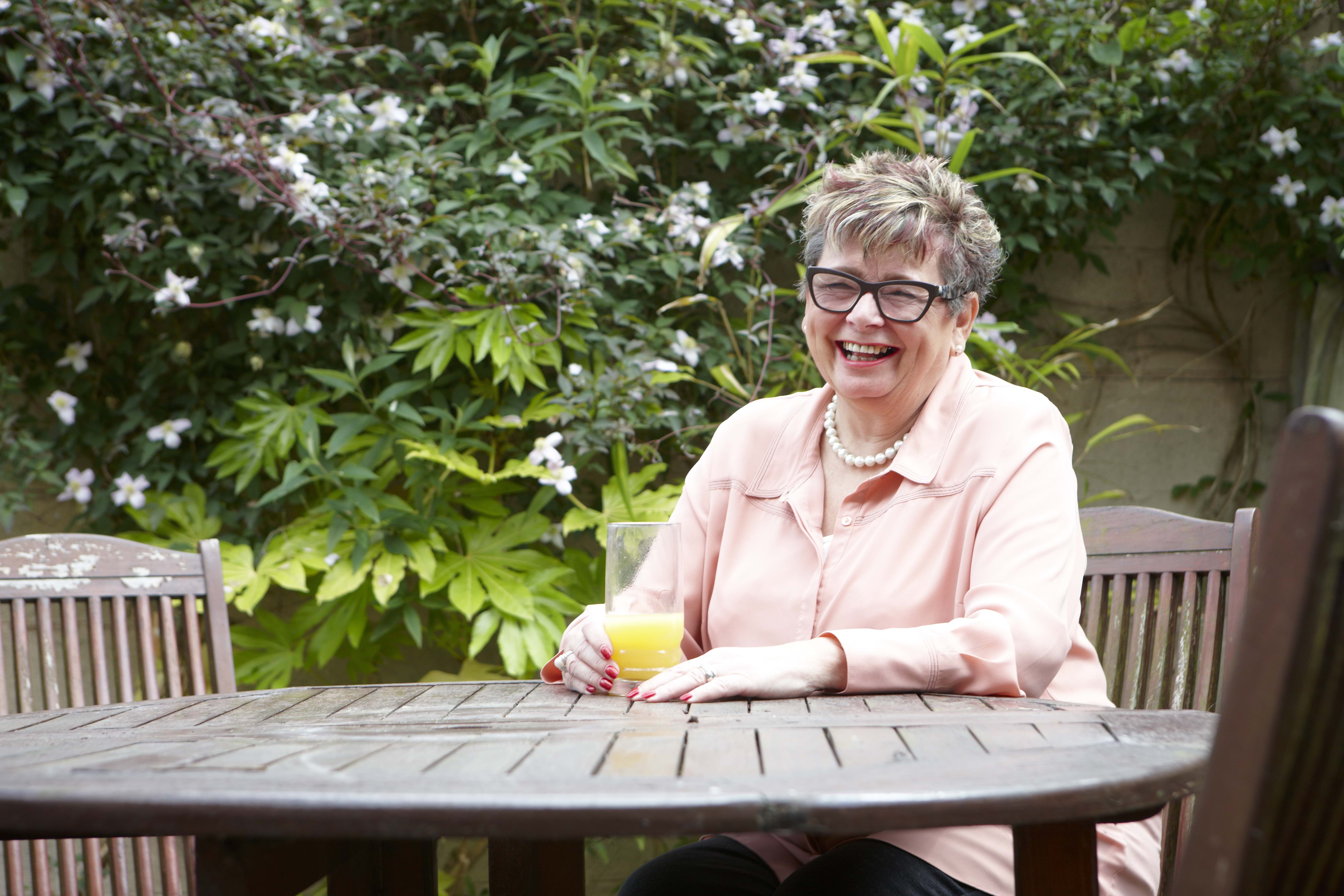 An elderly lady sits at a garden table smiling