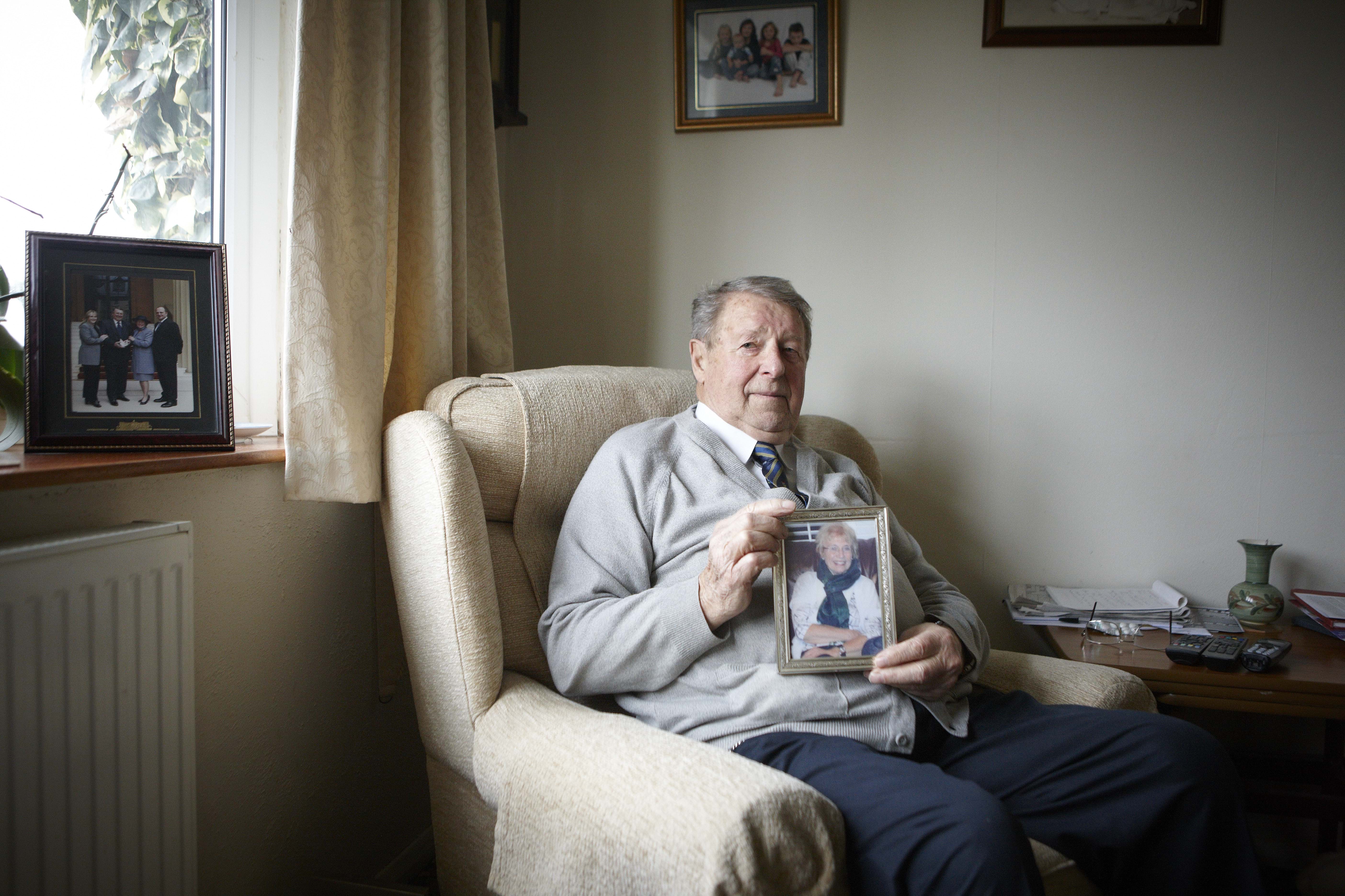 A man, sat in his living room, holds up an image of his wife, who passed away because of exposure to abestos