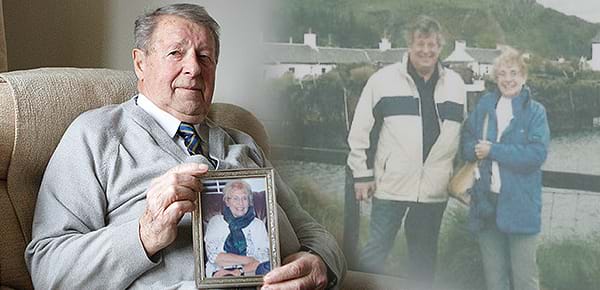 An elderly man sits in a chair holding an image of his wife