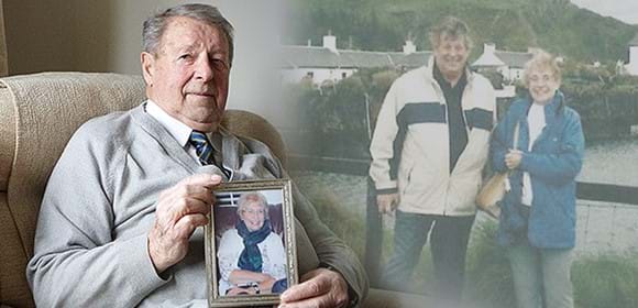 An elderly man sits in a chair holding an image of his wife