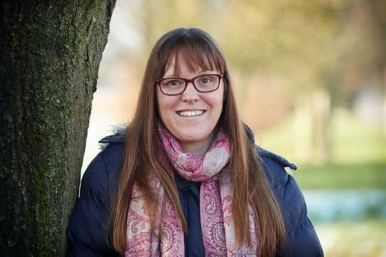 Margie Maguire standing next to a tree in a navy coat with a pink scarf.