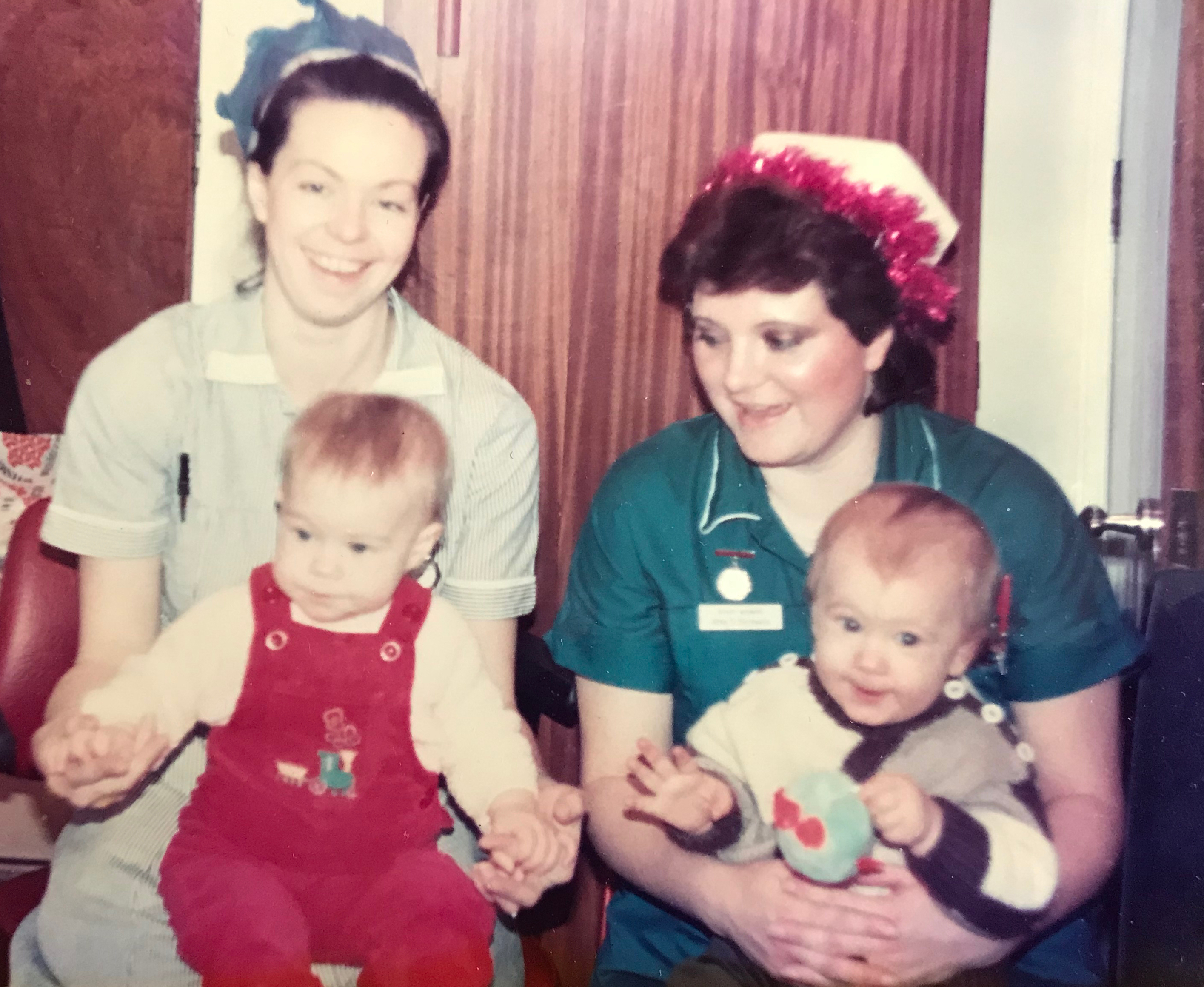 Two women dressed in medical uniforms hold two young children on a sofa