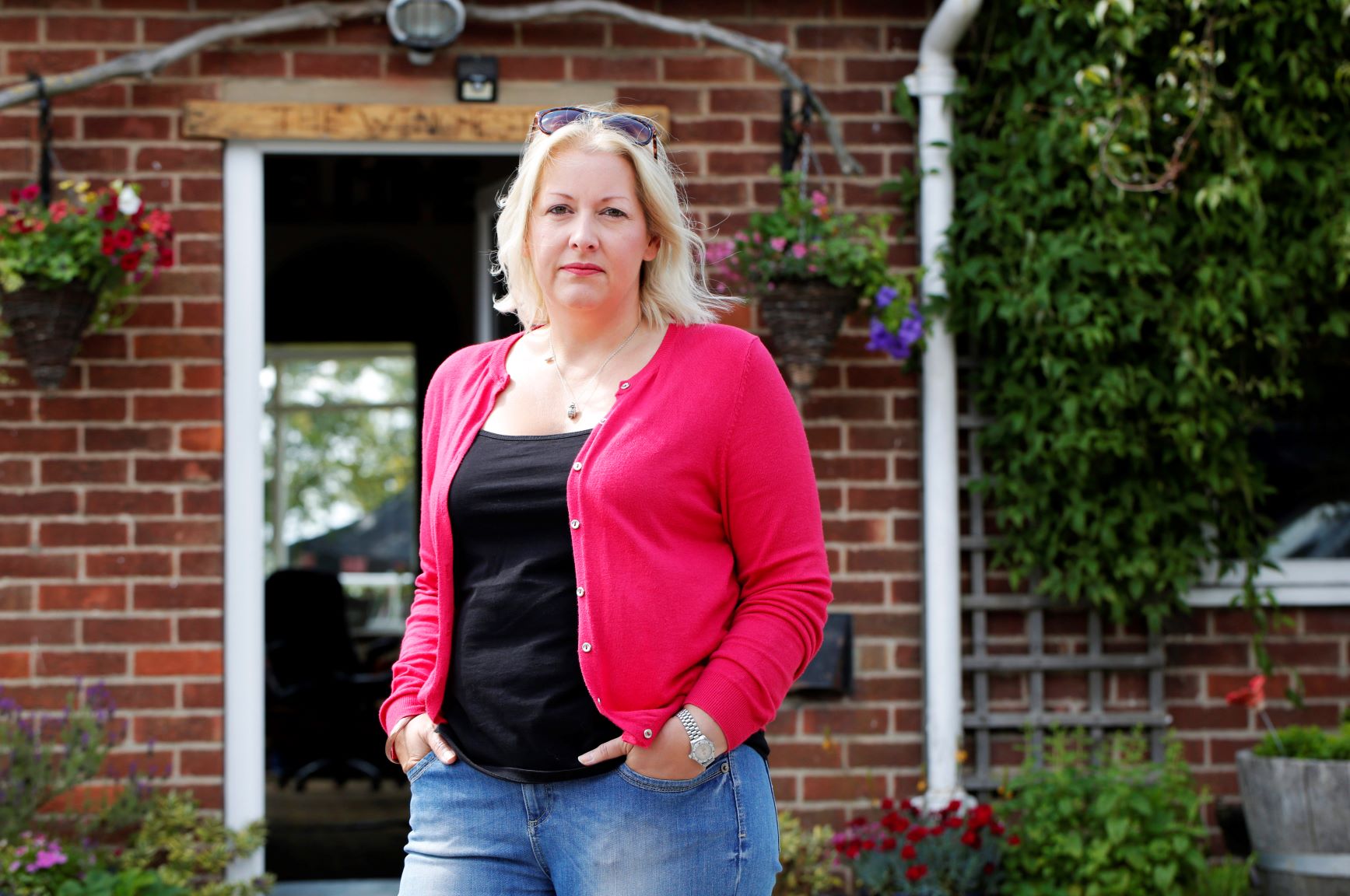 A blonde lady in a pink cardigan and black top stands with her hands in her pockets